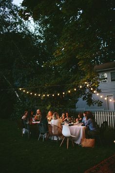 a group of people sitting around a dinner table in the backyard at night with string lights