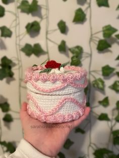 a hand holding a crocheted pink and white box with red flower on top