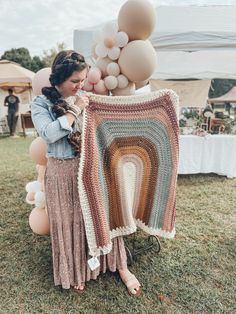 a woman is holding up a crocheted blanket in the grass with balloons behind her