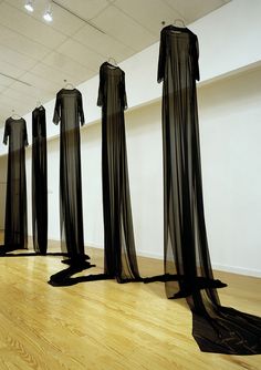 four black sheer dresses are on display in an empty room with wooden floors and white walls