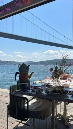 a table that has some food on top of it near the water with a bridge in the background