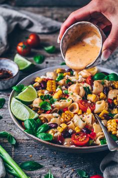 a person pouring dressing into a bowl filled with pasta and veggies on a wooden table