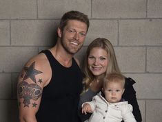 a man, woman and baby standing in front of a brick wall with tattoos on their arms