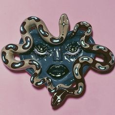 a blue and brown mask with large snakes on it's face, against a pink background