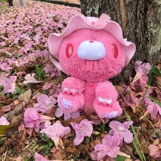 a pink teddy bear sitting on the ground next to a tree with purple flowers around it