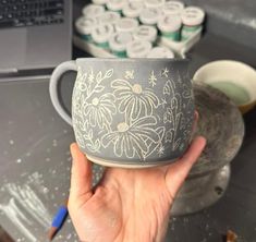 a hand holding a cup with flowers painted on it next to other cups and saucers