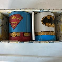 two coffee mugs with superman and batman symbol on them in a cardboard box next to each other