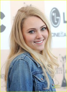 a woman with long blonde hair and blue denim jacket smiling at the camera while standing in front of a yellow background