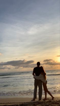 a man and woman standing on the beach at sunset with their arms around each other