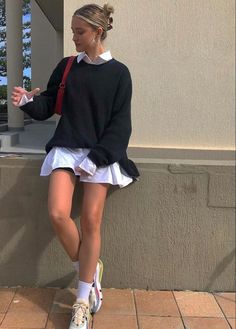 a woman sitting on the side of a building wearing a black sweater and white skirt