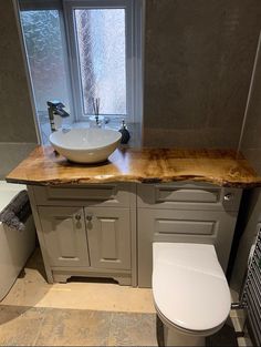 a white toilet sitting next to a sink in a bathroom under a window with a wooden counter top