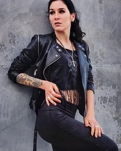 a woman leaning against a wall with her hand on her hip, wearing black pants and a leather jacket