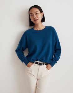 a woman standing in front of a white wall wearing a blue sweater and cream pants