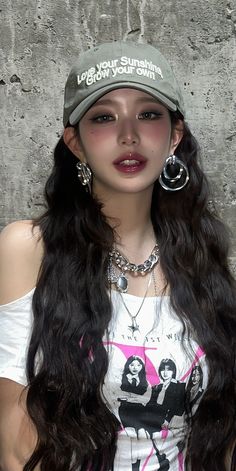 a woman with long hair wearing a baseball cap and earring necklaces standing in front of a concrete wall