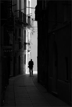 black and white photograph of man walking down an alleyway in the middle of town