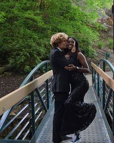 two people are hugging on a bridge in the woods, one is wearing a black dress