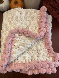a pink and white crocheted blanket sitting on top of a wooden table next to pumpkins