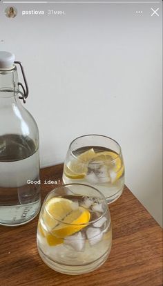 two glasses filled with ice and lemon on top of a wooden table next to a jug