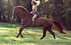 a woman riding on the back of a brown horse through a lush green forest filled field