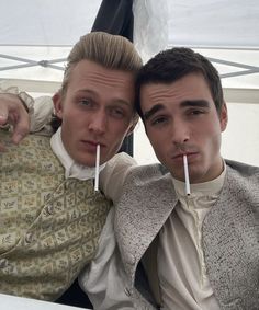 two men sitting next to each other with sticks sticking out of their mouths in front of them