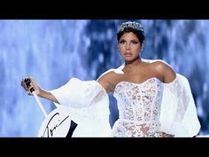 a woman wearing a white dress and holding an umbrella in her right hand while standing on stage