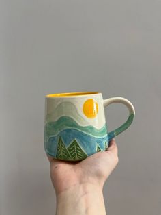 a hand holding a coffee mug with mountains and trees painted on the inside, in front of a gray background