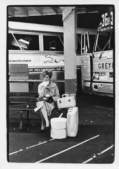 black and white photograph of woman sitting on bench with suitcases next to busses