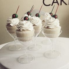 four cupcakes with white frosting and cherries in wine glasses on a cake stand