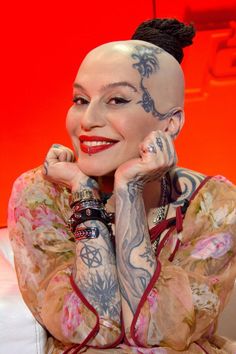 a woman with tattoos on her face and arm sitting in front of a red wall