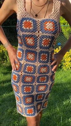 a woman standing in the grass wearing a crocheted dress with orange and blue squares on it