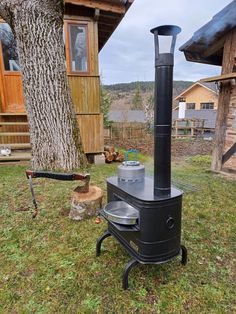 an outdoor wood burning stove in the grass next to a tree and shed with logs