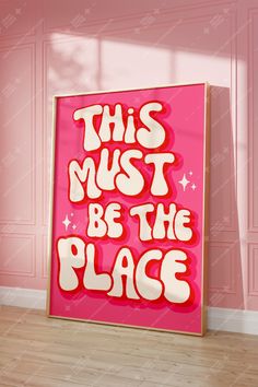 this must be the place sign in front of a pink wall with white lettering on it