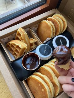 a person is holding a donut in a box with other pastries and condiments