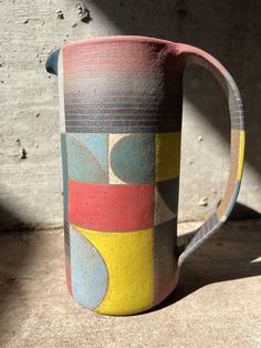 a multicolored ceramic pitcher sitting on the ground next to a wall with sunlight coming through it