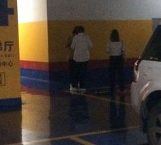 two people standing in front of a white car at a gas station with yellow and blue stripes on the wall