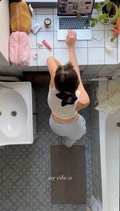 a woman standing in front of a laptop computer on top of a bathroom counter next to a sink