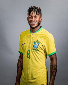 a man with dreadlocks standing in front of a gray background wearing a yellow shirt