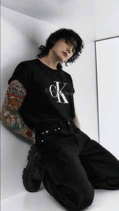 a man with tattoos sitting on the floor wearing black pants and a t - shirt