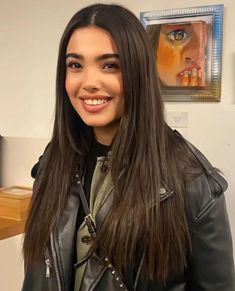 a woman with long hair wearing a black leather jacket and smiling at the camera while standing in front of a painting