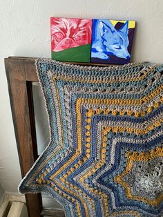 a crocheted blanket sitting on top of a wooden chair next to two paintings