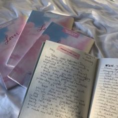 three notebooks with writing on them are laying on a bed sheet and next to an open book