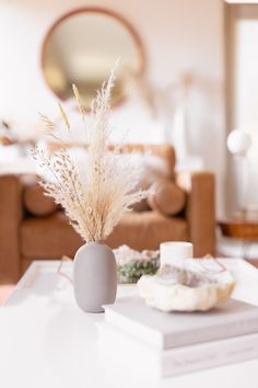 a white table topped with a gray vase filled with dry grass next to a brown couch