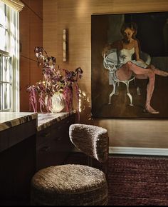 a painting hangs on the wall next to a chair and vase with flowers in it