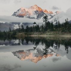 the mountains are covered in clouds and trees as they reflect in the still lake water