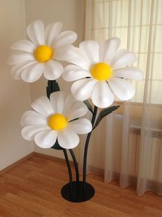 three white flowers in a black vase on a wooden floor next to a window with sheer curtains