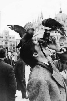 a man holding a camera up to his face with two birds perched on top of him