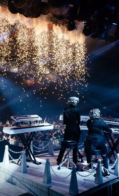 two people sitting at a table watching fireworks go off in the sky behind them on stage