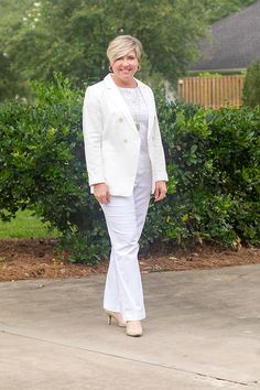 a woman standing in front of bushes wearing a white suit and heels with her hands in her pockets