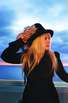 a woman with long blonde hair wearing a black hat and feathered jacket on the beach