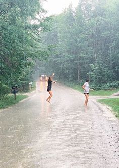 two people running in the rain on a road with trees behind them and one person holding an umbrella